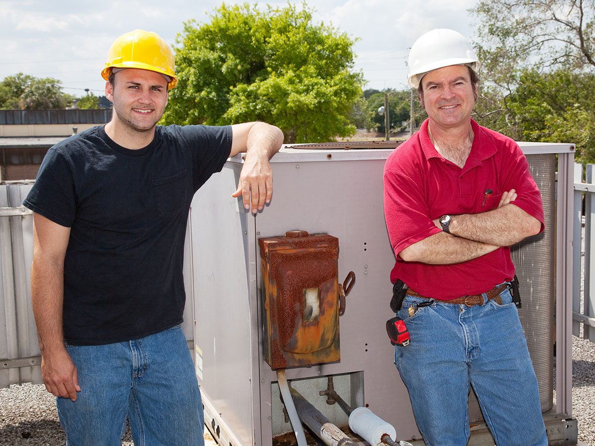 Two men wearing hard hats, leaning up against an HVAC unit and smiling.